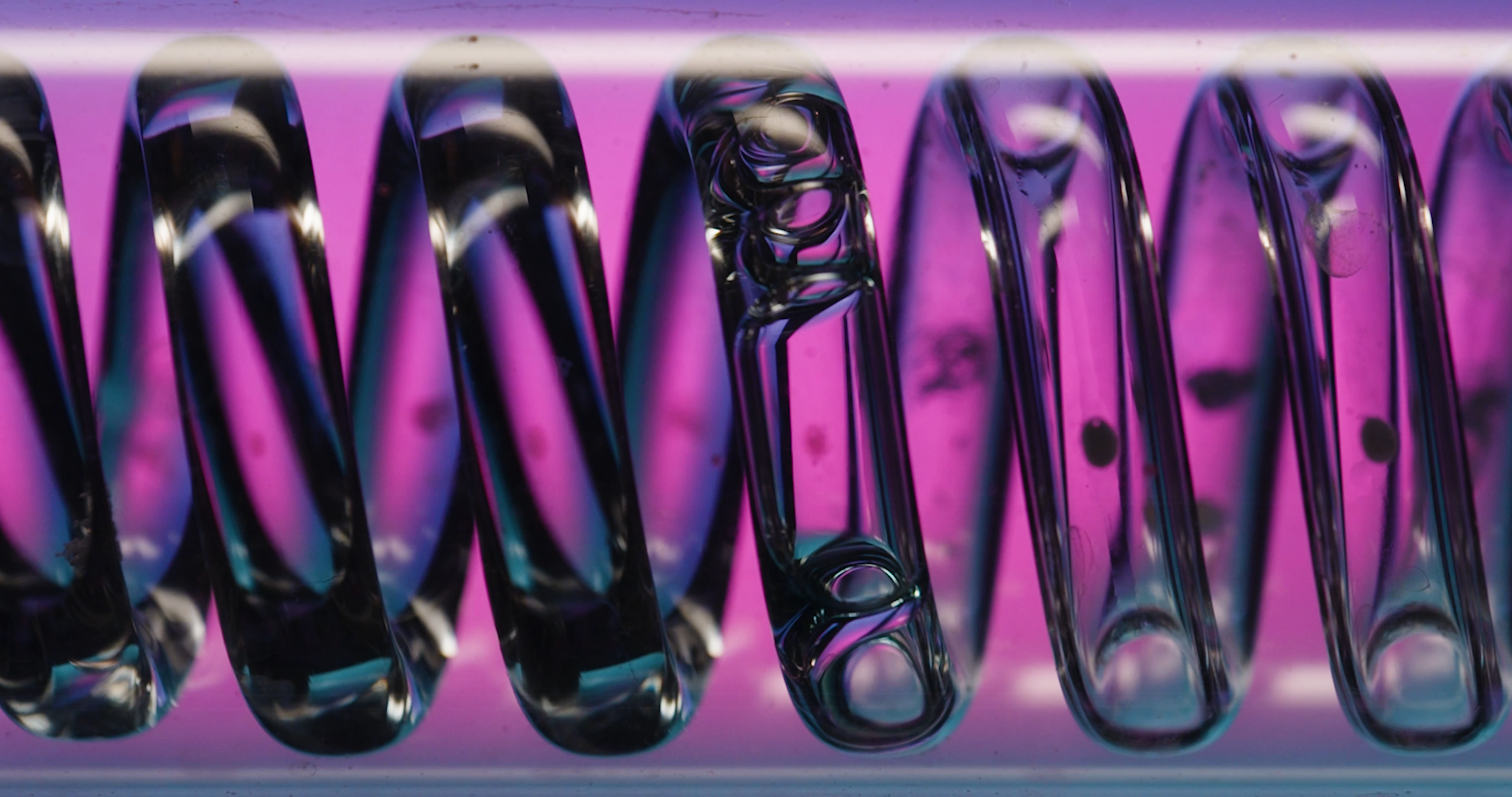 A spiral-shaped tube with fluid flowing through it is seen against a pink backdrop in a lab setting, taken from our Bruker collaboration on Flow NMR